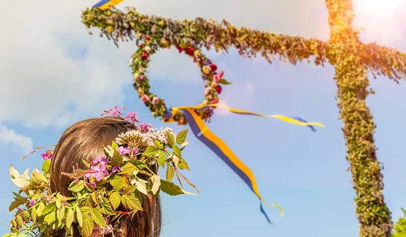 Midsommarklädd tjej som tittar upp på midsommarstång och en blå himmel
