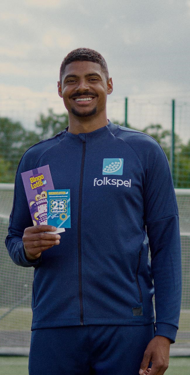 Michel Tornéus står framför ett fotbollsmål med Folkspels lotter BingoLotto och Sverigelotten i handen